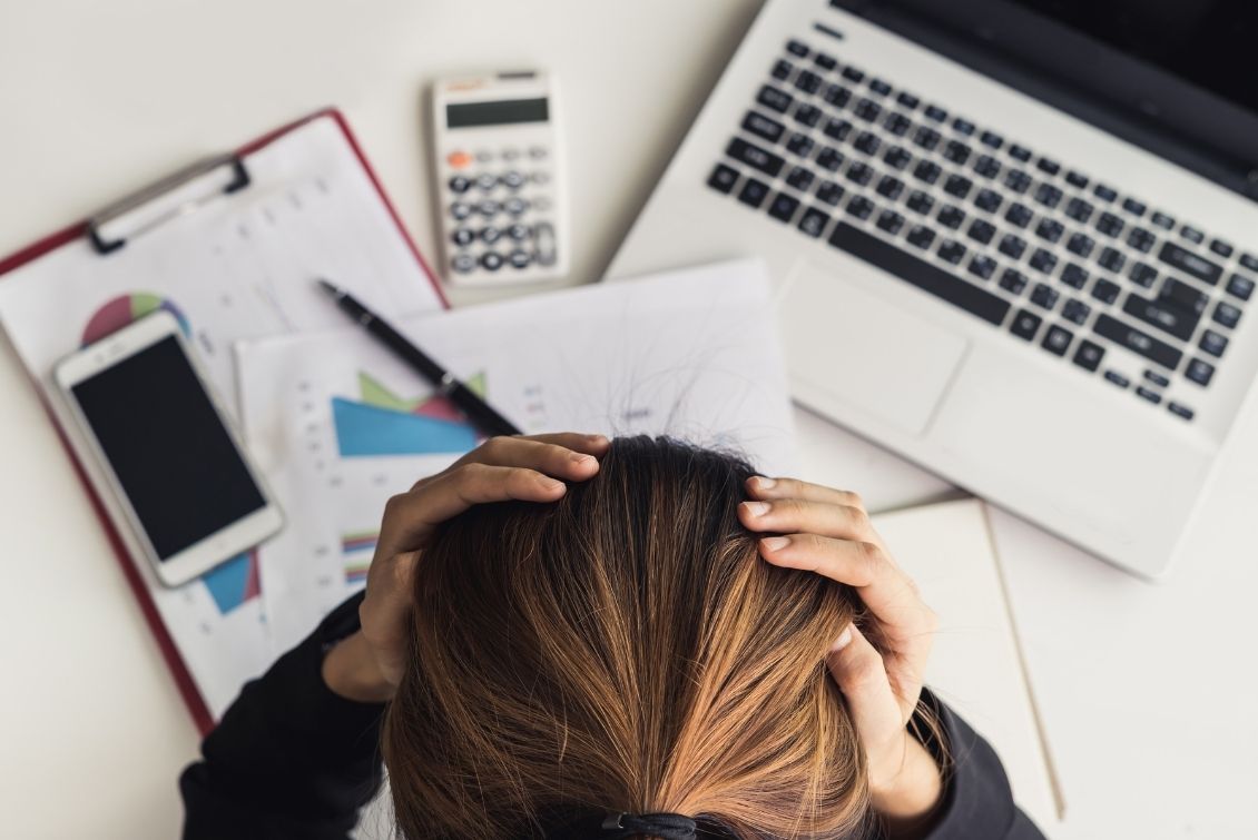 A woman has her hands on her head as she looks down at papers, a calculator and a laptop