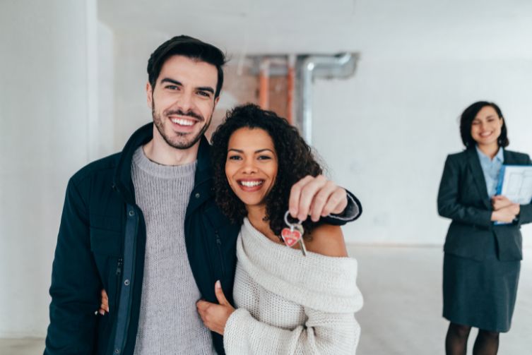 A young man and young woman hold each other as he holds up a house key with a smiling realtor in the background